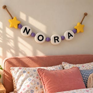 May include: A wall-mounted name banner featuring the letters N, O, R, and A printed in black on large white circular beads. The banner is strung on a thick rope with yellow star-shaped accents at each end and small purple beads.