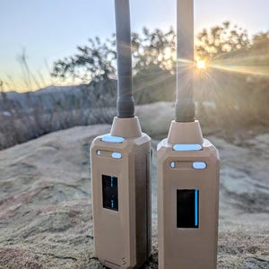 May include: Two tan, rectangular handheld radio transceivers with vertical antennas. Each device features a small, dark digital display screen, two light-coloured rectangular buttons near the top, and a small logo at the bottom of the casing.