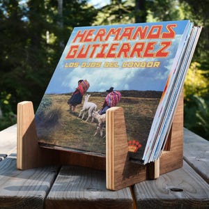 May include: A wooden vinyl record display stand holding a record album titled Hermanos Gutierrez Los Ojos Del Condor. The album cover features two people in traditional clothing walking with two llamas in a grassy, mountainous landscape.