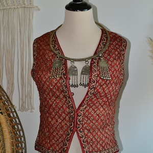 Puede incluir: Un chaleco acolchado rojo con estampado floral y un collar plateado con colgantes. El chaleco tiene cierre de botón y ribete negro.