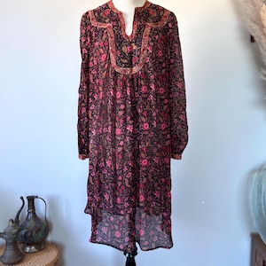 May include: A long-sleeved, knee-length dress with a floral pattern in shades of pink and brown. The dress has a V-neckline with decorative trim and is displayed on a black dress form. The dress is made of a sheer fabric.