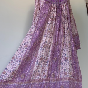 Könnte beinhalten: Ein langes, fließendes Kleid mit floralem Muster in Lila-, Weiß- und Beigetönen. Das Kleid hat lange Ärmel, einen hohen Kragen und eine dekorative Bordüre am Saum. Das Kleid wird auf einer Schaufensterpuppe präsentiert.