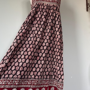 Puede incluir: Un vestido largo sin mangas con un estampado floral burdeos y crema. El vestido presenta un escote cuadrado y tirantes anchos. El dobladillo y el escote están adornados con un borde decorativo. El vestido se exhibe en un maniquí.
