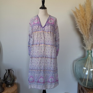 Puede incluir: Vestido de manga larga con estampado floral en blanco, morado y rosa, expuesto en un maniquí. El vestido tiene cuello en V y detalles dorados. El vestido, que llega a la rodilla, tiene un corte holgado y fluido.