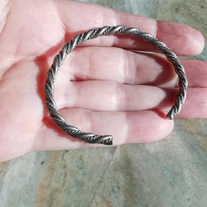 Puede incluir: Un brazalete de metal retorcido de color plateado. El brazalete tiene un diseño similar a una cuerda y está abierto en los extremos. El brazalete descansa sobre una superficie de color claro.