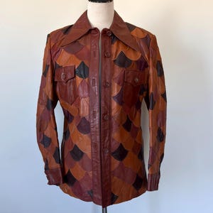 May include: A vintage leather jacket with a unique patchwork design. The jacket features a button-up front, two chest pockets, and a collar. The color palette includes shades of brown, tan, and black, creating a striking visual texture. The jacket is displayed on a mannequin.