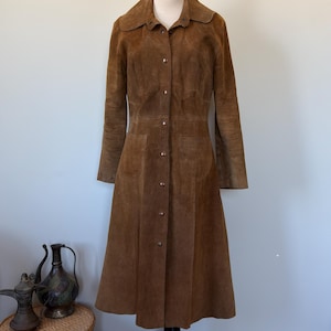 May include: A vintage brown suede coat with a button-down front, long sleeves, and a flared skirt. The coat has a collar and two front pockets. It is displayed on a black mannequin. The coat is approximately 55 inches long.