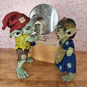 May include: Two green goblin figurines facing each other. One wears a red hat, yellow shirt, and brown pants, cleaning a round mirror. The other wears a blue dress and tan apron, with a tan headscarf, appearing to shout at the first goblin.