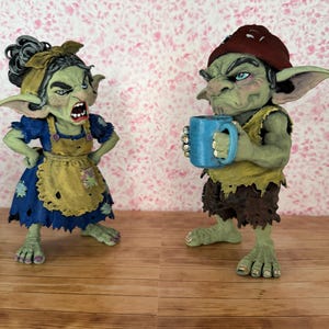 May include: Two green-skinned goblin figurines. The left figure wears a blue dress with a yellow apron and a yellow headband. The right figure wears a yellow top, brown shorts, and a red beanie, holding a blue mug with both hands.