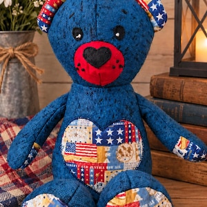 May include: A stuffed bear made of dark blue textured fabric with a red muzzle and black heart-shaped nose. The bear features patches on its ears, feet, and a large heart on its chest, all displaying patriotic American flag and liberty-themed prints.