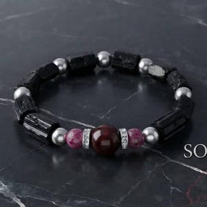 Könnte beinhalten: Ein Perlenarmband mit zylindrischen schwarzen Steinen, silberfarbenen Kugeln, zwei kleineren rosa gesprenkelten Perlen und einer zentralen dunkelroten Kugel, die von zwei silberfarbenen Ringen mit klaren Steinen flankiert wird. Text SOLENNE S.
