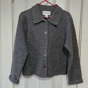 May include: A charcoal gray long-sleeve jacket with a textured finish, featuring a classic collar, five dark buttons down the front, and a David Hollis brand label on the inside neck. The jacket has straight sleeves with folded cuffs at the ends.