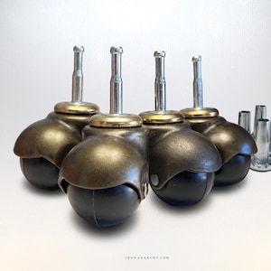 May include: A set of four furniture casters with dark bronze-toned metal hoods, black spherical wheels, and silver-colored metal stems. Three separate silver-colored metal mounting sockets are positioned to the right of the casters.