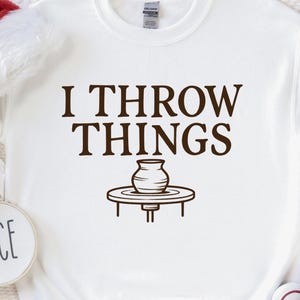 May include: A white long-sleeve sweatshirt featuring the brown text I THROW THINGS above a graphic of a pottery wheel and vase. A red and white Santa hat, two small circular ornaments labeled NAUGHTY and NICE, and white sneakers are present.