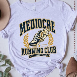 May include: A white short-sleeve t-shirt featuring a graphic of a winged running shoe in yellow and navy blue. The text reads MEDIOCRE RUNNING CLUB EST. EVENTUALLY in navy blue and yellow block lettering, accompanied by yellow laurel leaf accents.
