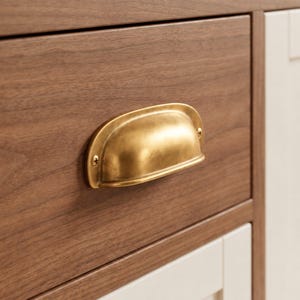 May include: A brass cup pull handle with a brushed finish attached to a dark brown wood drawer front. The hardware features a curved, semi-circular design with two visible screws securing it to the wooden surface of the furniture piece.