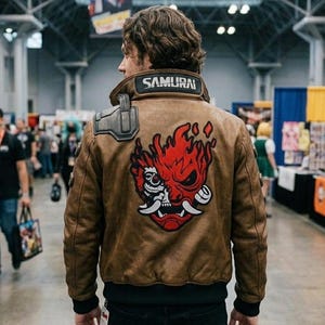 Könnte beinhalten: Eine braune Leder-Bomberjacke mit einer großen rot-weißen Oni-Masken-Grafik auf dem Rücken. Der Kragen hat einen schwarzen Aufnäher mit dem weißen Schriftzug SAMURAI. Sie verfügt über ein dunkelgraues Schulterteil und schwarze Rippbündchen.