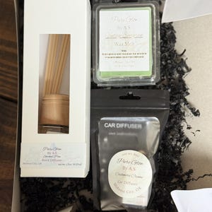 May include: A gift box containing a white box of Pure Glow By A.S. Smoked Pine reed diffuser, a clear plastic container of green Vintage Evergreen wax melt, and a black pouch of Pure Glow By A.S. Cashmere Creme car diffuser.