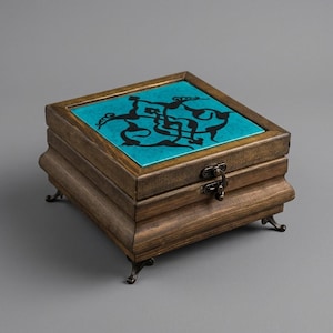 May include: A square wooden box with a dark brown finish, featuring a bright turquoise lid inlay decorated with a black swirling graphic pattern. The box stands on four small, curved metal feet and includes a central metal latch closure.