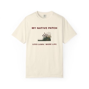 May include: A cream-colored short-sleeve t-shirt featuring the dark red text MY NATIVE PATCH and LESS LAWN / MORE LIFE. Between the lines of text is a graphic illustration of a small patch of native wildflowers and tall grasses.
