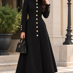 May include: A long black coat featuring a high collar, a full-length front button closure with gold-toned buttons, long sleeves with three gold-toned buttons at each cuff, and a structured, flared skirt silhouette, paired with a small black handbag.