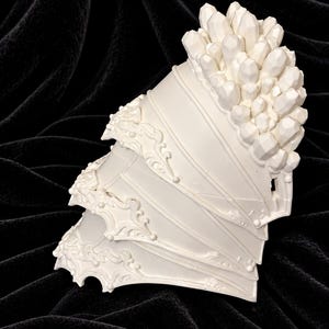 May include: A white shoulder armor piece featuring three layered plates with ornate, raised scrollwork along the edges and a cluster of faceted, crystal-like geometric protrusions extending from the top section of the armor.
