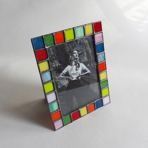 May include: A colorful picture frame with a black and white photo of a woman. The frame is made of glass tiles in shades of red, yellow, green, blue, and pink.