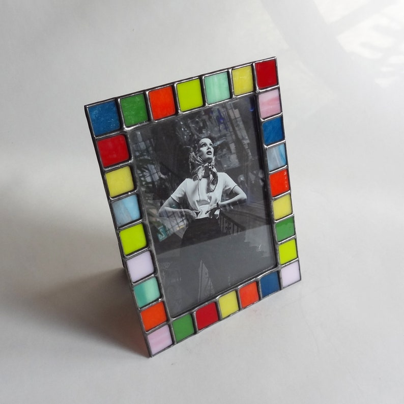 May include: A colorful picture frame with a black and white photo of a woman. The frame is made of glass tiles in shades of red, yellow, green, blue, and pink.