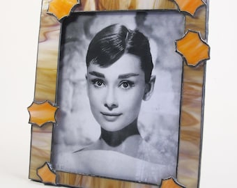 May include: A stained glass picture frame with a brown and orange design. The frame has a starburst pattern in the corners and a black and white photo of a woman inside.