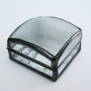 Op de afbeelding: Een kleine, vierkante glazen doos met een zilverkleurig metalen frame. De bovenkant van de doos is van helder glas met een textuurpatroon en kleine luchtbellen. De onderkant van de doos is van helder glas met een bloemenpatroon.