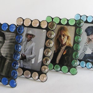 May include: Four decorative picture frames with different coloured glass beads around the edges. The frames are arranged in a row from left to right with the blue frame in the front, followed by the pink frame, the green frame, and the light blue frame at the back.