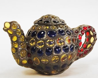 May include: A decorative teapot with a dark metal frame and colorful glass accents. The teapot features yellow, blue, and red glass pieces. The handle and spout are also adorned with the same colorful glass. The lid is also made of dark metal and glass.