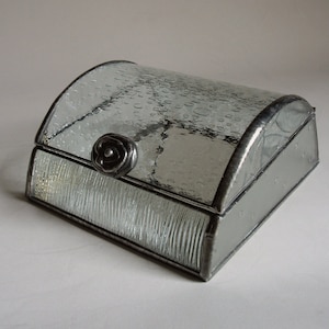 May include: A clear glass box with a textured lid and a metal rose-shaped knob. The box is made of stained glass and has a decorative design.