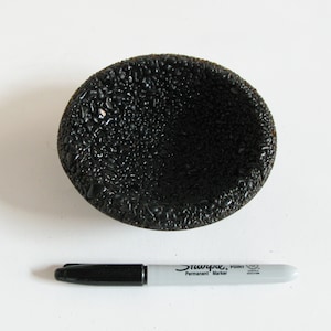 May include: A black, round, textured bowl with a slightly raised rim. The bowl is made of a material that resembles small, black pebbles.