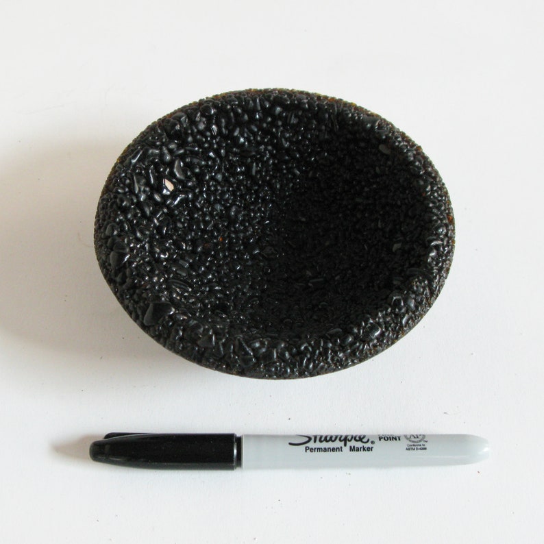 May include: A black, round, textured bowl with a slightly raised rim. The bowl is made of a material that resembles small, black pebbles.