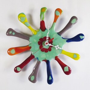 Op de afbeelding: Een abstracte wandklok met een gespat ontwerp. De klok heeft een lichtgroen midden met een rood bloemmotief en uitstralende armen in verschillende kleuren, waaronder geel, rood, blauw en grijs. De wijzers zijn wit en de secondewijzer is rood.
