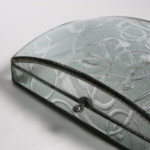 May include: A light blue glass jewelry box with a silver knob and a decorative etched design. The box has a curved top and a black metal frame.