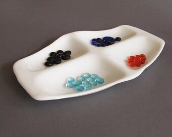 May include: A white ceramic divided tray with four distinct sections, each containing small, smooth, rounded glass stones in black, dark blue, bright orange, and light blue colors, arranged in a scattered pattern within the dish.