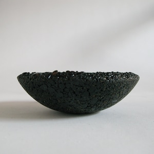 May include: A black ceramic bowl with a rough, textured surface. The bowl is shaped like a half-sphere and has a slightly uneven rim.