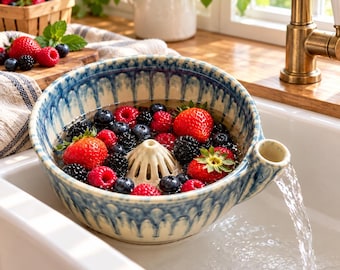 Könnte beinhalten: Ein Beeren-Seiher aus Keramik mit blau-cremefarbener Tropfglasur, einem zentralen Kegel und einem seitlichen Ausguss. Die Schale ist mit frischen Erdbeeren, Himbeeren und Brombeeren gefüllt, die in einem weißen Spülbecken abgespült werden.