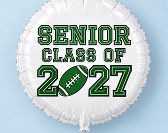 May include: A round white foil balloon featuring the dark green text SENIOR CLASS OF 2027. The number zero in the year 2027 is replaced by a green and white American football graphic. The balloon has a metallic sheen and a white ribbon attached.