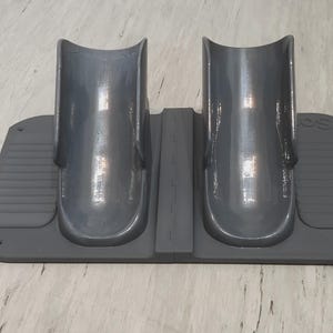 May include: A dark gray plastic foot alignment device featuring two curved, upright cradles mounted on a rectangular base. The base includes textured side panels with horizontal ridges and a central divider, designed to support and position feet.