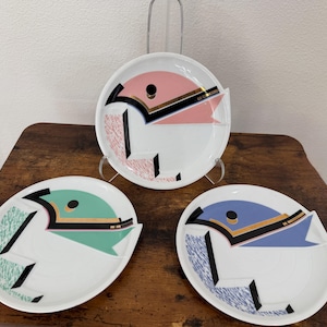 Puede incluir: Tres platos de cerámica blanca con diseños gráficos geométricos. Cada plato muestra una forma estilizada de ave en rosa, verde o azul, acentuada con negro, oro y patrones texturizados. Un plato se exhibe verticalmente en un soporte.