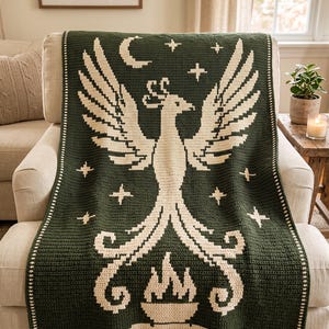 Könnte beinhalten: Eine dunkelgrüne Häkeldecke mit einem großen cremefarbenen Phönix, der aus Flammen aufsteigt. Das Design zeigt eine Mondsichel und mehrere vierzackige Sterne um den Vogel herum. Die Decke hat einen einfachen cremefarbenen Rand.