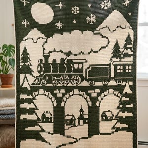 May include: A rectangular crochet blanket featuring a dark forest green and cream color scheme. The design depicts a steam train traveling across a stone bridge above a small village, with mountains, a large moon, and snowflakes in the night sky.