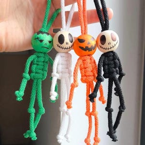 May include: Four macrame ornaments featuring wooden bead heads with painted faces. The figures are green, white, orange, and black, each with a matching cord loop for hanging. The bodies are constructed from knotted cord with simple limbs.