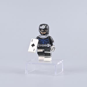 May include: A plastic minifigure character wearing a black mask with a white target symbol on the forehead, a black suit with blue muscle detailing, and white accents. The figure holds a white playing card featuring a black ace of spades symbol.