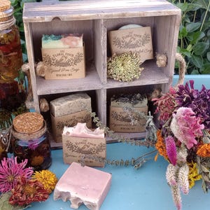 May include: A collection of handmade soap bars with paper labels reading The Floral Finch, three glass jars filled with dried flowers and cork stoppers, and a bouquet of dried wildflowers arranged on a light blue wooden tray.