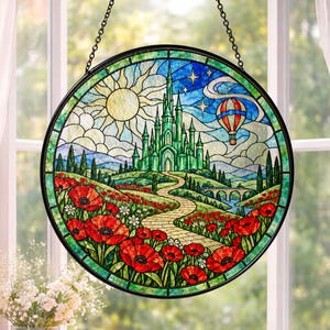 Puede incluir: Un atrapasueños circular de vidrieras con un castillo verde, un sol amarillo y un globo aerostático en un cielo azul. Un camino de ladrillos amarillos serpentea a través de un campo de amapolas rojas y flores blancas hacia el castillo.