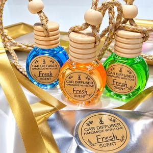 May include: Three round glass car diffuser bottles filled with blue, orange, and green liquid. Each bottle has a light wood cap, a hanging rope, and a circular label reading CAR DIFFUSER HANDMADE WITH LOVE Fresh SCENT.